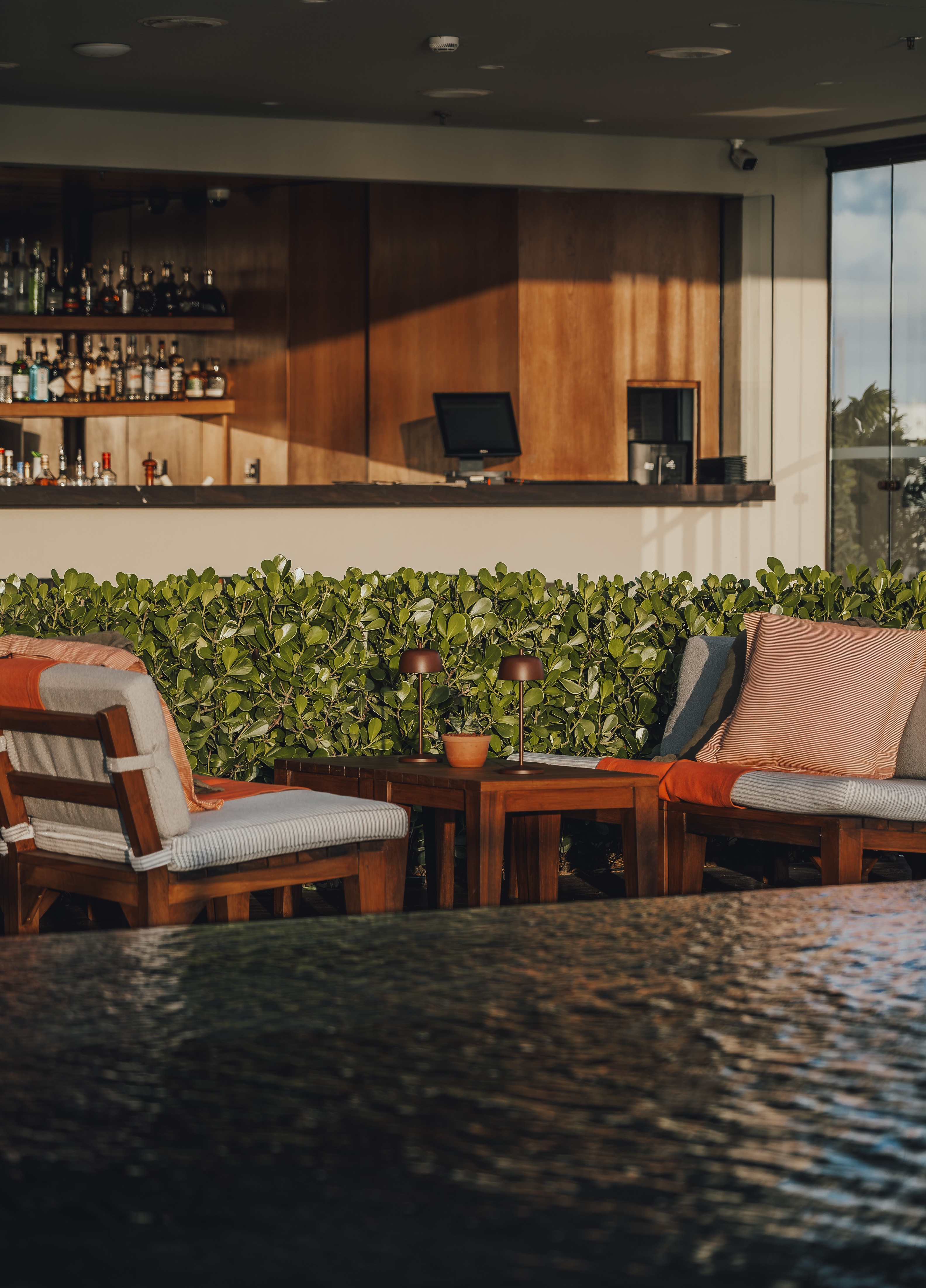 Warm-lit luxury hotel bar lounge with teak seating, linen cushions, green hedge wall, and fully stocked back bar at Fasano